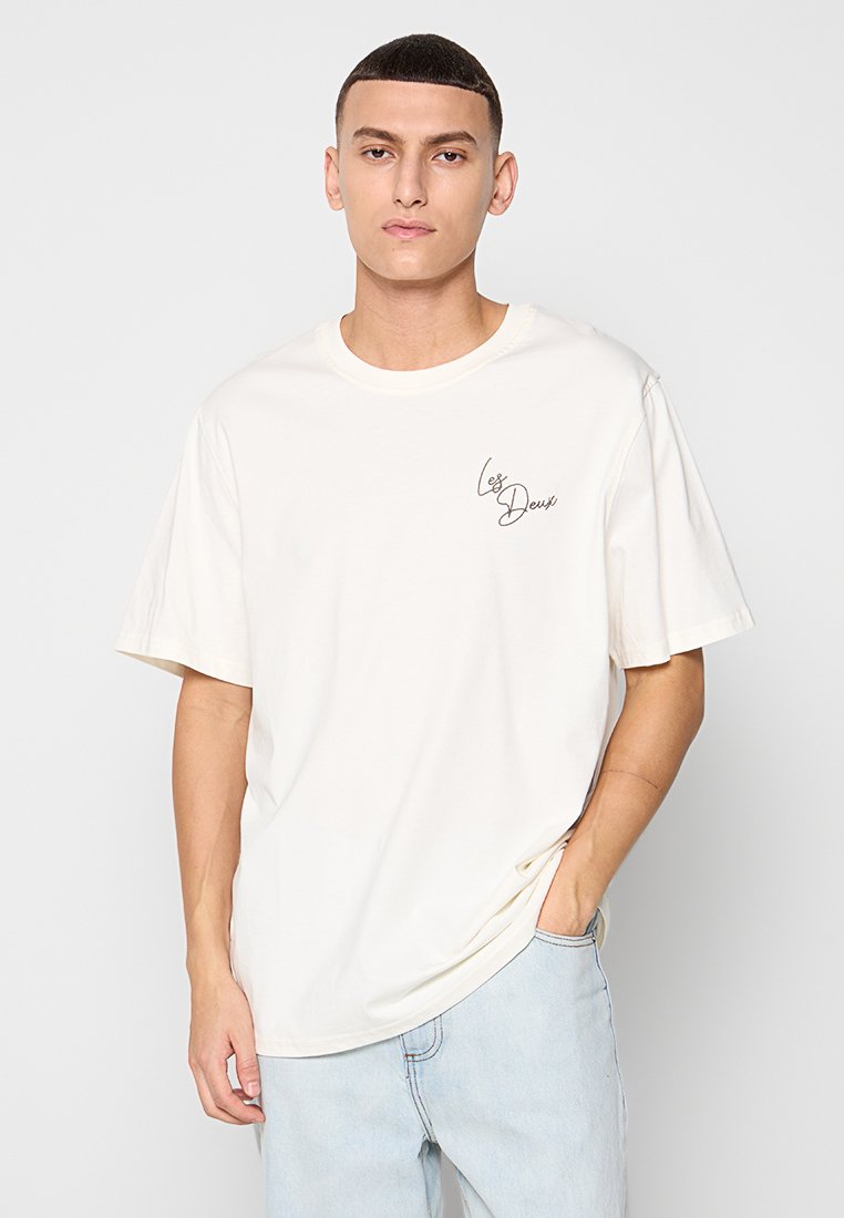 Cremfarbene Baumwoll-T-Shirt mit lockerer Passform, kurzen Ärmeln, Rundhalsausschnitt und einem kleinen schwarzen Schriftzug-Logo auf der linken Brust.