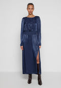 Marineblaues Kleid mit langen Ärmeln, geraffter Taille, seitlichem Schlitz und glatter, glänzender Textur. Getragen mit braunen kniehohen Stiefeln.