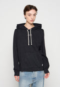 Junge Person mit kurzen braunen Haaren, die einen schwarzen Hoodie mit weißen Kordeln und hellblauen Jeans vor einfarbigem Hintergrund trägt.