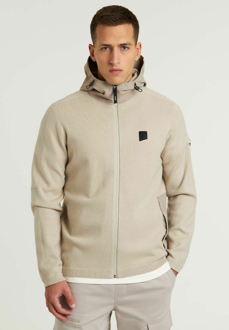 Beige Zip-Jacke mit Kapuze aus strukturiertem Stoff. Verfügt über ein schwarzes Logo auf der Brust und zwei seitliche Taschen. Einfaches, funktionales Design.