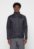 Mann trägt eine schwarze gesteppte Reißverschlussjacke und graue Hose, steht vor einem einfarbigen hellen Hintergrund und schaut nach vorne.