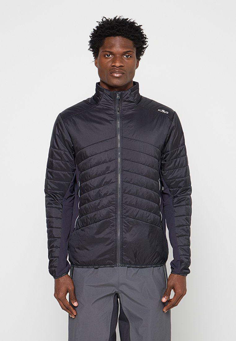 Mann trägt eine schwarze gesteppte Reißverschlussjacke und graue Hose, steht vor einem einfarbigen hellen Hintergrund und schaut nach vorne.