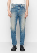 Mann in abgetragenen, schmal geschnittenen blauen Jeans, weißem Hemd und schwarzen Slippern, der vor einem weißen Hintergrund steht.