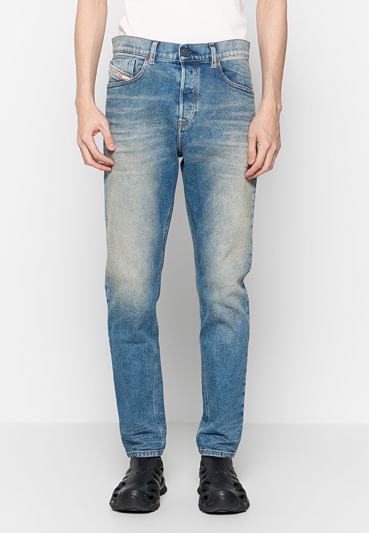 Mann in abgetragenen, schmal geschnittenen blauen Jeans, weißem Hemd und schwarzen Slippern, der vor einem weißen Hintergrund steht.