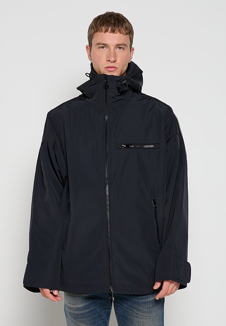 Junger Mann in schwarzer Kapuzenjacke mit Reißverschlusstaschen und blauen Jeans, der vor einem einfarbigen hellen Hintergrund steht.