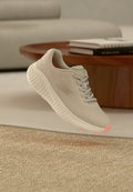 Beiger Sportschuh mit Mesh-Obermaterial und strukturierter Sohle schwebt über einem carpetbedeckten Boden in einem Wohnzimmer mit Büchern auf einem runden Holztisch.