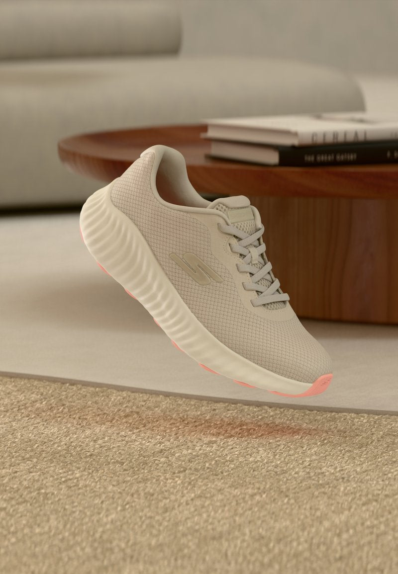 Beiger Sportschuh mit Mesh-Obermaterial und strukturierter Sohle schwebt über einem carpetbedeckten Boden in einem Wohnzimmer mit Büchern auf einem runden Holztisch.