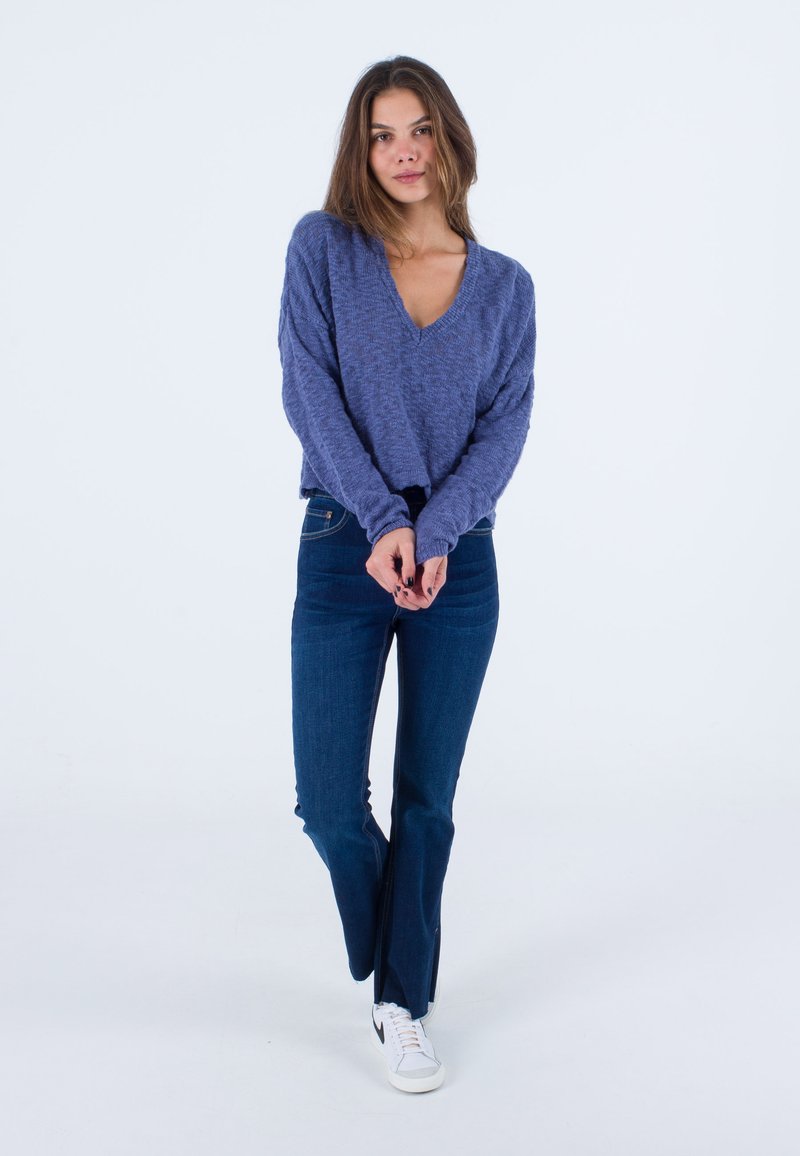 Lila Strickpullover mit V-Ausschnitt, weiter Passform und langen Ärmeln, kombiniert mit dunkelblauen Schlagjeans und weißen Turnschuhen.