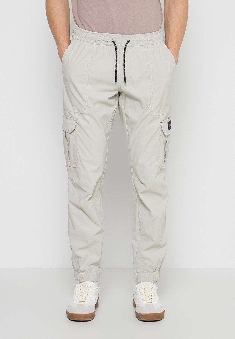 Mann trägt hellbeige Cargohosen mit elastischen Bündchen, schwarzem Kordelzug, Seitentaschen, kombiniert mit weißen Sneakern und einem hellbraunen Hemd.