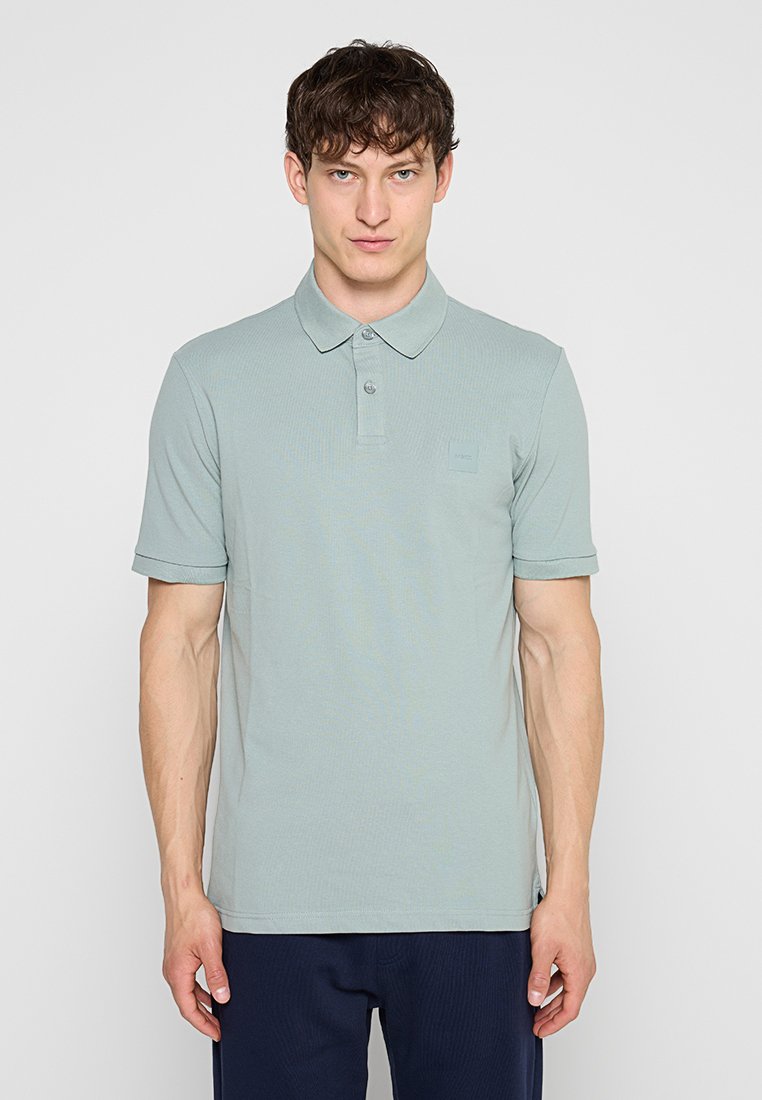 Junger Mann trägt ein hellgrünes Poloshirt mit kurzen Ärmeln und dunkle Hose, steht vor einem schlichten hellen Hintergrund.