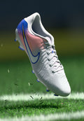 Weiße und rosa Nike Fußballschuhe mit blauen Details, die Gras auf einem markierten grünen Fußballfeld aufwirbeln.