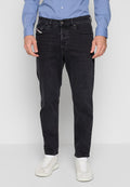 Mann trägt schmal geschnittene schwarze Jeans, ein hellblaues Hemd mit Knöpfen und weiße Sneaker, steht vor einem einfarbigen hellgrauen Hintergrund.