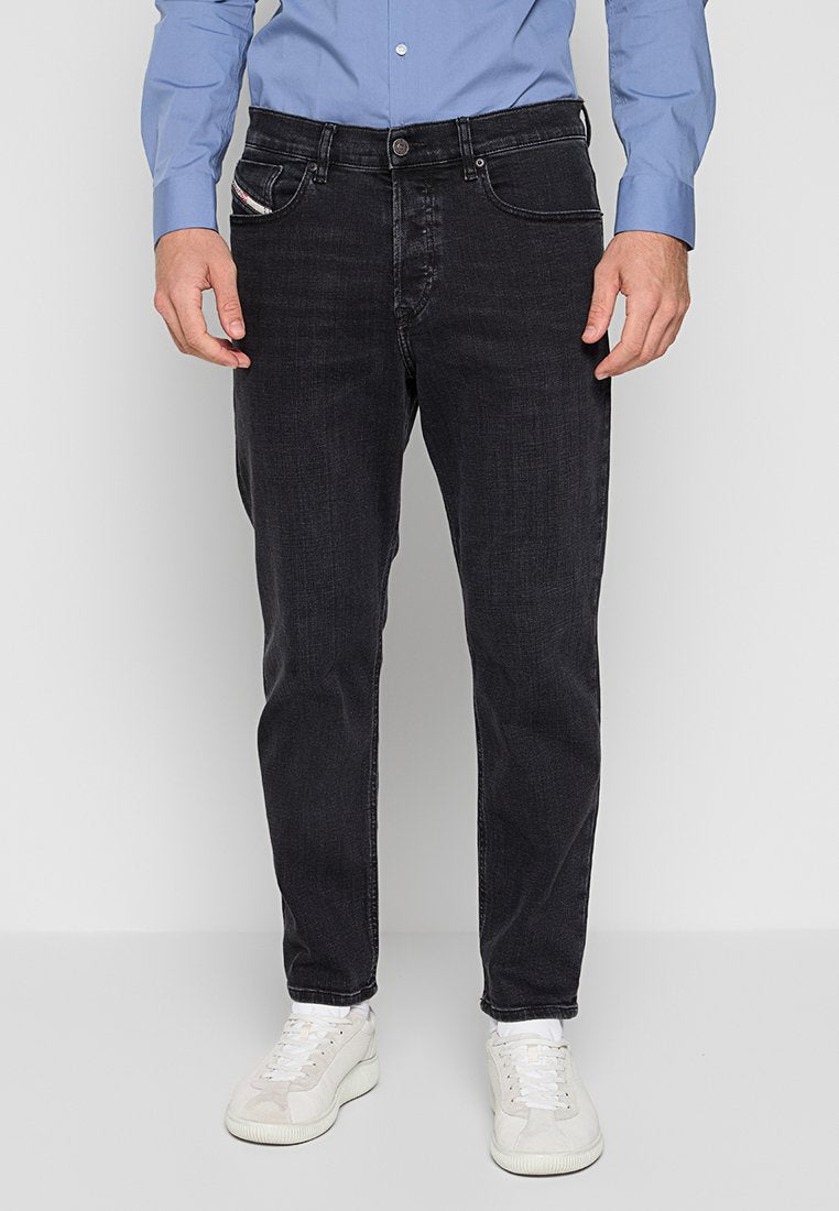 Mann trägt schmal geschnittene schwarze Jeans, ein hellblaues Hemd mit Knöpfen und weiße Sneaker, steht vor einem einfarbigen hellgrauen Hintergrund.