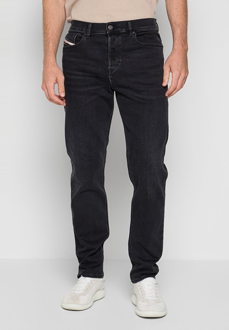 Mann in schwarzen, geraden Jeans und weißen Sneakers, der vor einem schlichten hellgrauen Hintergrund steht.
