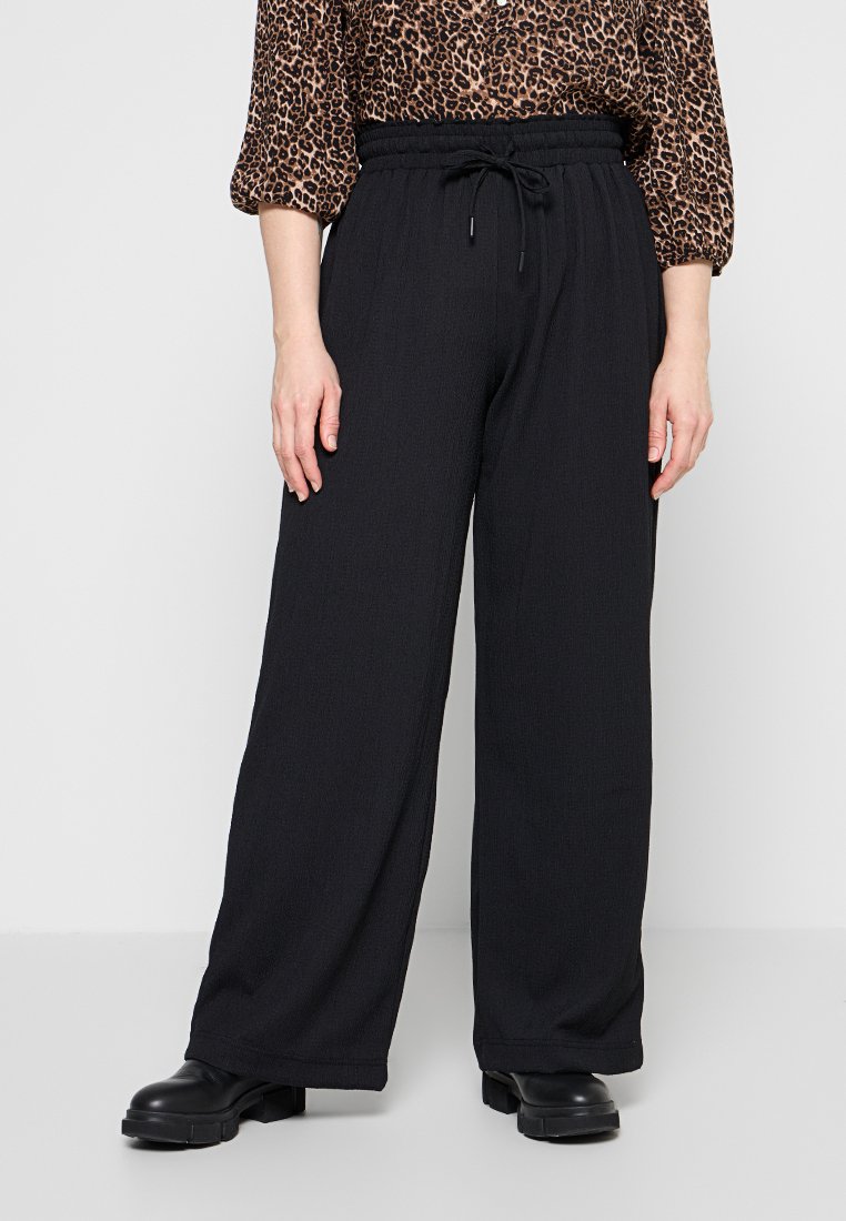 Frau trägt lockere schwarze Kordelzughose, Leopardenmuster-Bluse mit Puffärmeln und schwarze Stiefel mit klobiger Sohle, steht vor einer schlichten Wand.