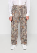Mann trägt beige Tarnhosen mit Baum- und Blattmustern, weiße Turnschuhe und ein weißes Langarmshirt, stehend vor einem schlichten Hintergrund.