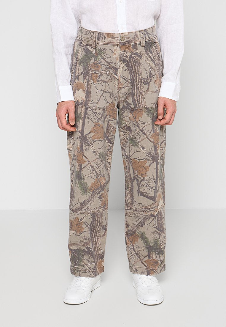 Mann trägt beige Tarnhosen mit Baum- und Blattmustern, weiße Turnschuhe und ein weißes Langarmshirt, stehend vor einem schlichten Hintergrund.