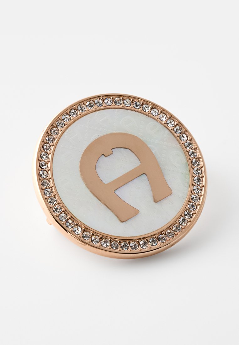 Runde Brosche mit roségoldener Umrandung, verziert mit klaren Kristallen, weißem Emaille-Zentrum und einem roségoldenen stilisierten Buchstaben „a“ in der Mitte.