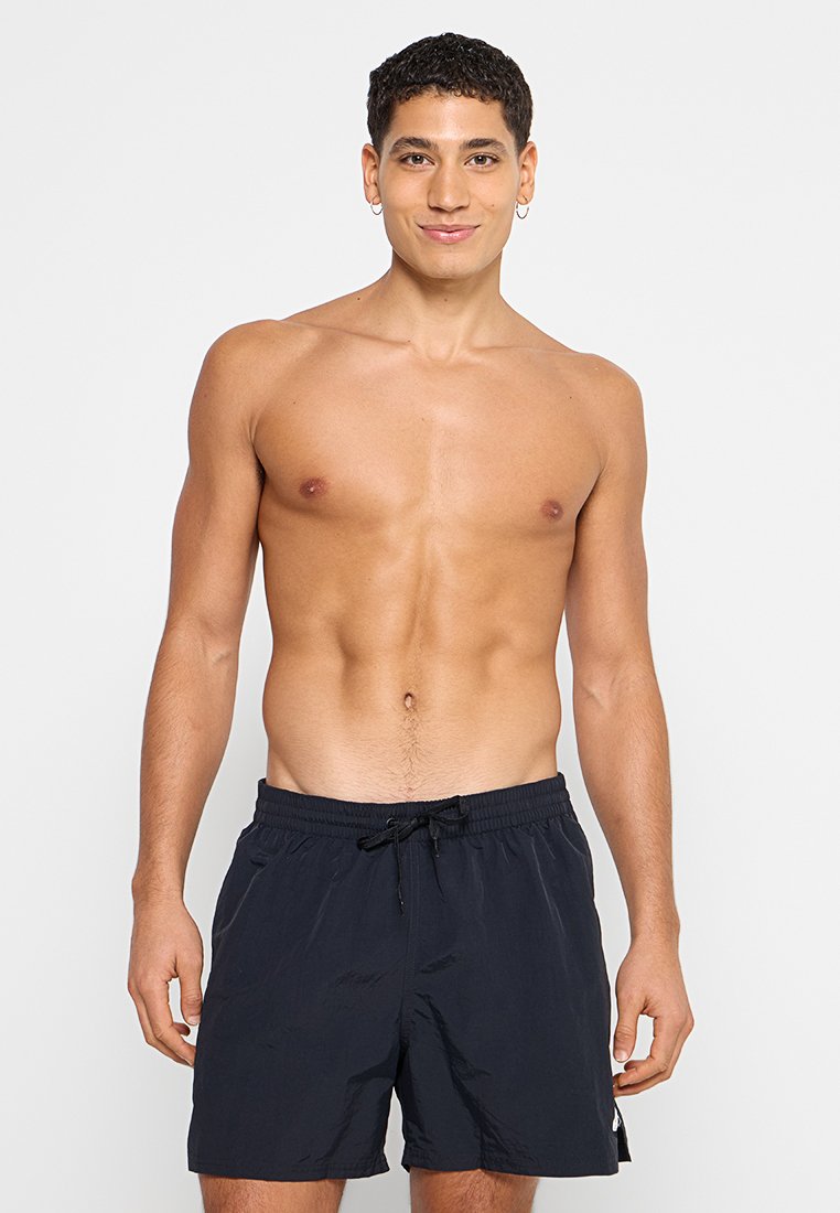 Junger Mann mit kurzen, lockigen Haaren, der schwarze Badeshorts trägt, stehend und lächelnd vor einem schlichten hellen Hintergrund.