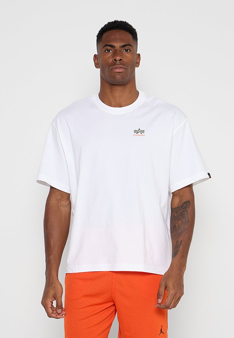 Mann mit kurzen Haaren, der ein weißes übergroßes T-Shirt mit einem kleinen Logo und leuchtend orangefarbene, perforierte Shorts trägt und vor einem schlichten Hintergrund steht.