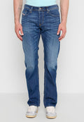 Mann trägt blaue Jeans mit geradem Bein und vertikalen Verblassungslinien, kombiniert mit weißen und grauen Sneakers, steht vor einem schlichten Hintergrund.