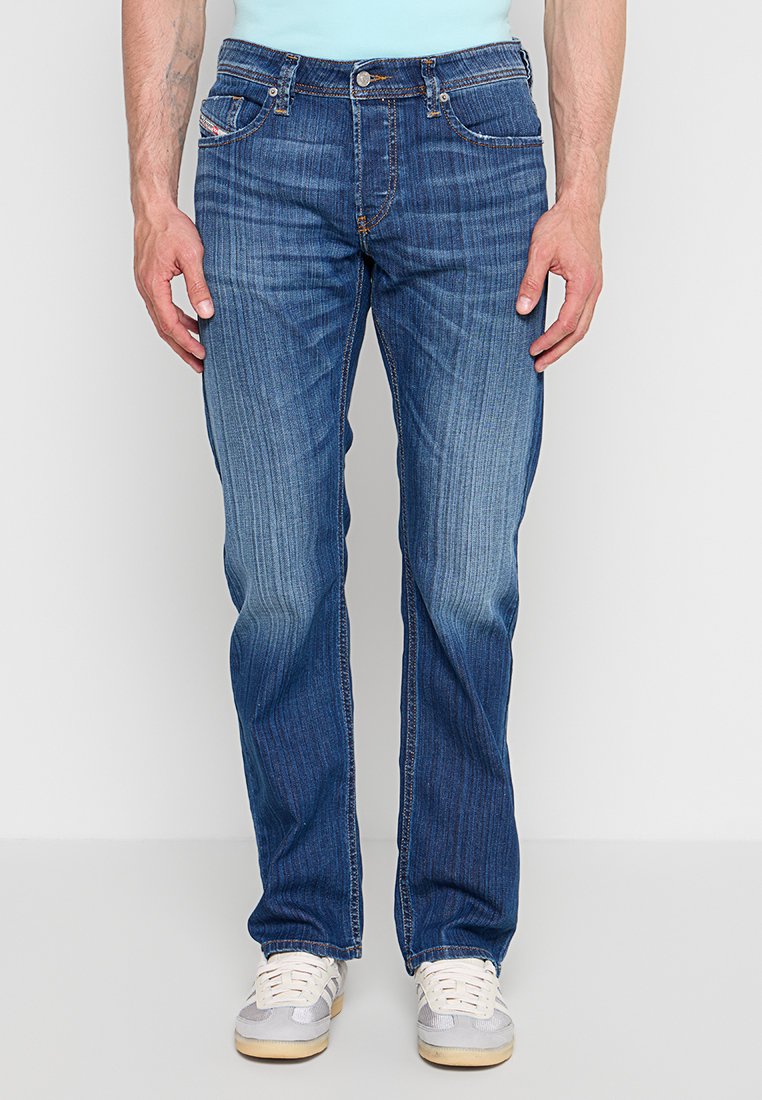 Mann trägt blaue Jeans mit geradem Bein und vertikalen Verblassungslinien, kombiniert mit weißen und grauen Sneakers, steht vor einem schlichten Hintergrund.
