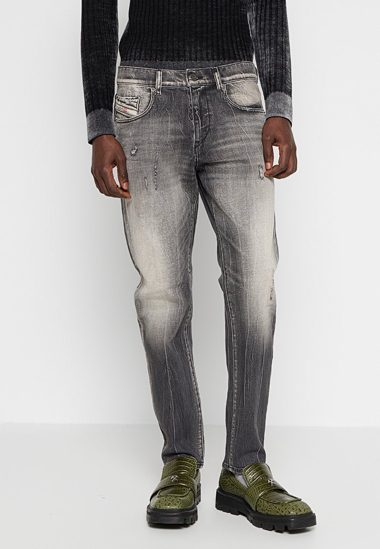 Schwarzer gestrickter Pullover mit geripptem Muster, kombiniert mit grauen, ausgewaschenen Jeans, die sichtbare Abnutzungsspuren aufweisen. Grüne strukturierten Schuhe.