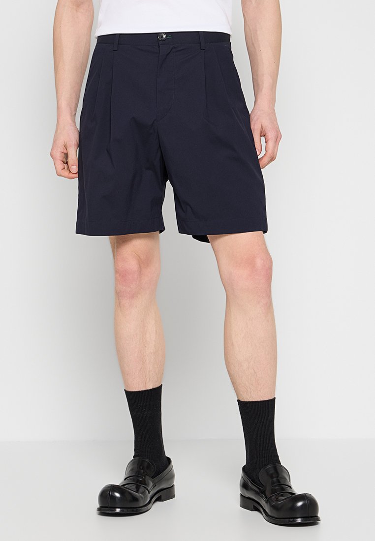 Dunkelblaue Shorts mit vorderen Falten, einem Knopfverschluss, schrägen Seitentaschen und einer taillierten Passform. Kombiniert mit schwarzen Socken und glänzenden Loafers.