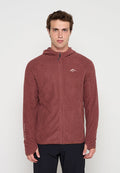 Junger Mann trägt einen weinroten Fleecepullover mit Reißverschluss und schwarze Hose, stehend aufrecht vor einem einfarbigen hellen Hintergrund.