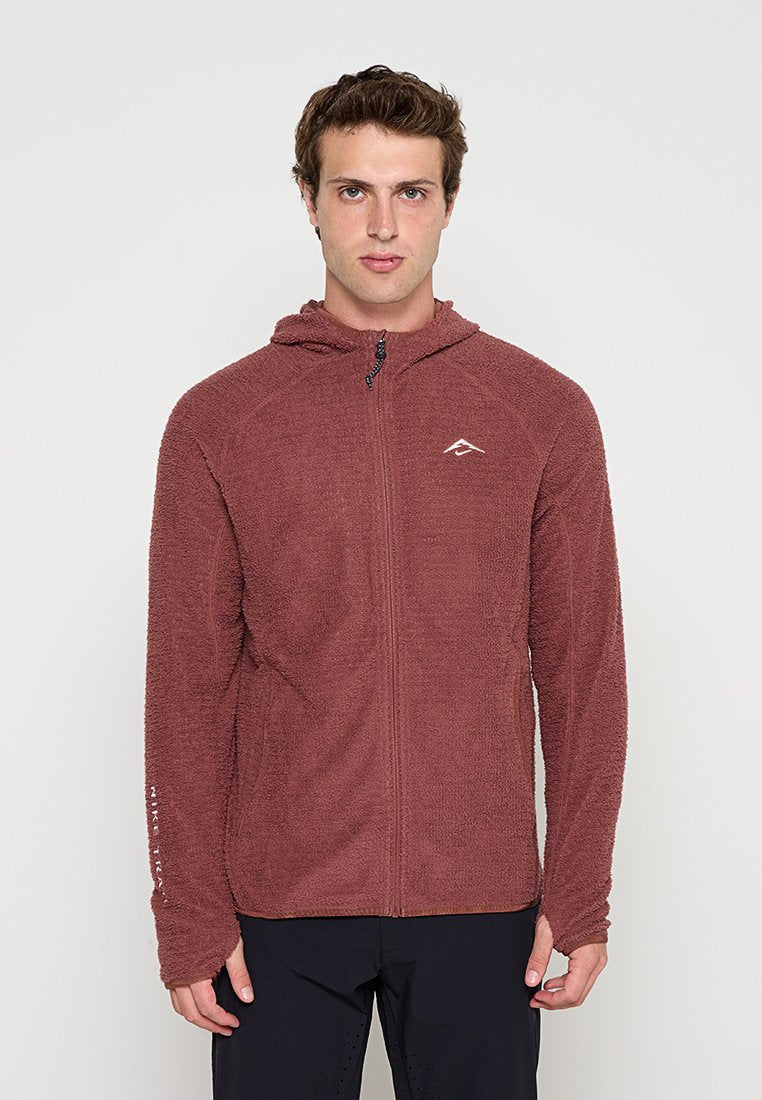 Junger Mann trägt einen weinroten Fleecepullover mit Reißverschluss und schwarze Hose, stehend aufrecht vor einem einfarbigen hellen Hintergrund.