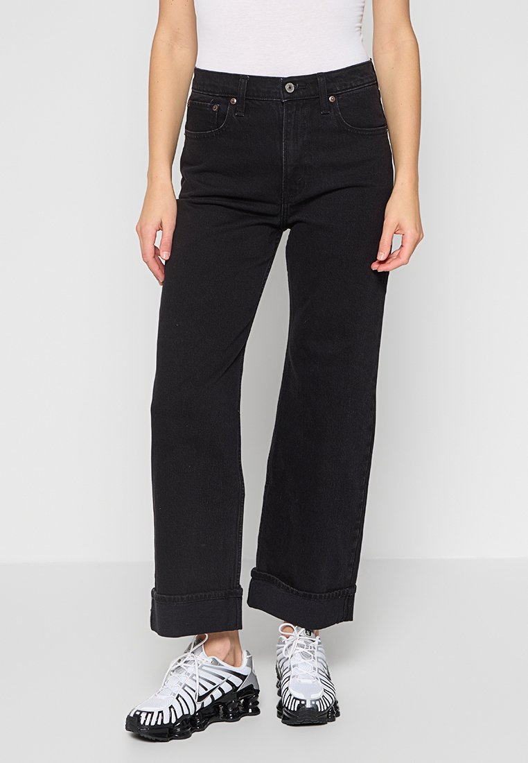 Schwarze hochtaillierte weit geschnittene Jeans aus Denim, mit einem Knopfverschluss und umgeschlagenen Säumen, kombiniert mit schwarz-weißen gemusterten Turnschuhen.