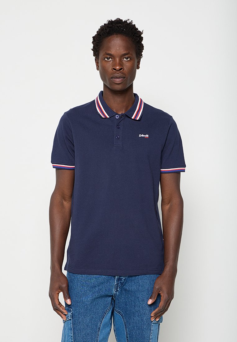 Mann trägt ein marineblaues Poloshirt mit einem rot-weiß-blau gestreiften Kragen und Ärmelabschlüssen, kombiniert mit blauen Jeans aus Denim.