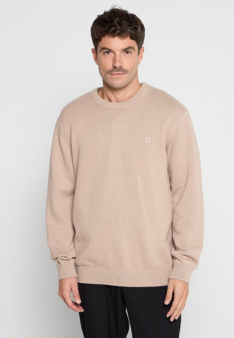 Beige Strickpullover mit strukturiertem Rippdesign, Rundhalsausschnitt, langen Ärmeln und einem kleinen Logo auf der Brust, kombiniert mit dunklen Hosen.