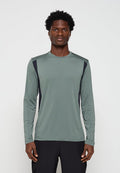 Junger Mann mit kurzen lockigen Haaren, der ein langärmeliges grünes Sportshirt mit schwarzen Seitenteilen trägt, steht vor einem schlichten Hintergrund.