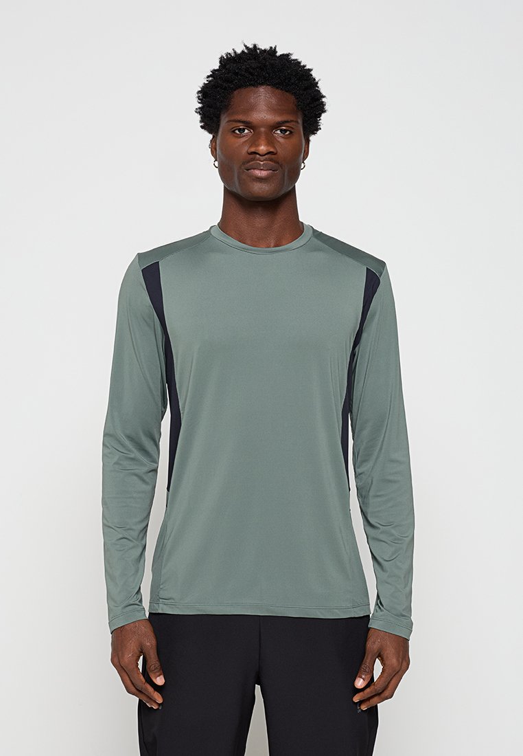 Junger Mann mit kurzen lockigen Haaren, der ein langärmeliges grünes Sportshirt mit schwarzen Seitenteilen trägt, steht vor einem schlichten Hintergrund.