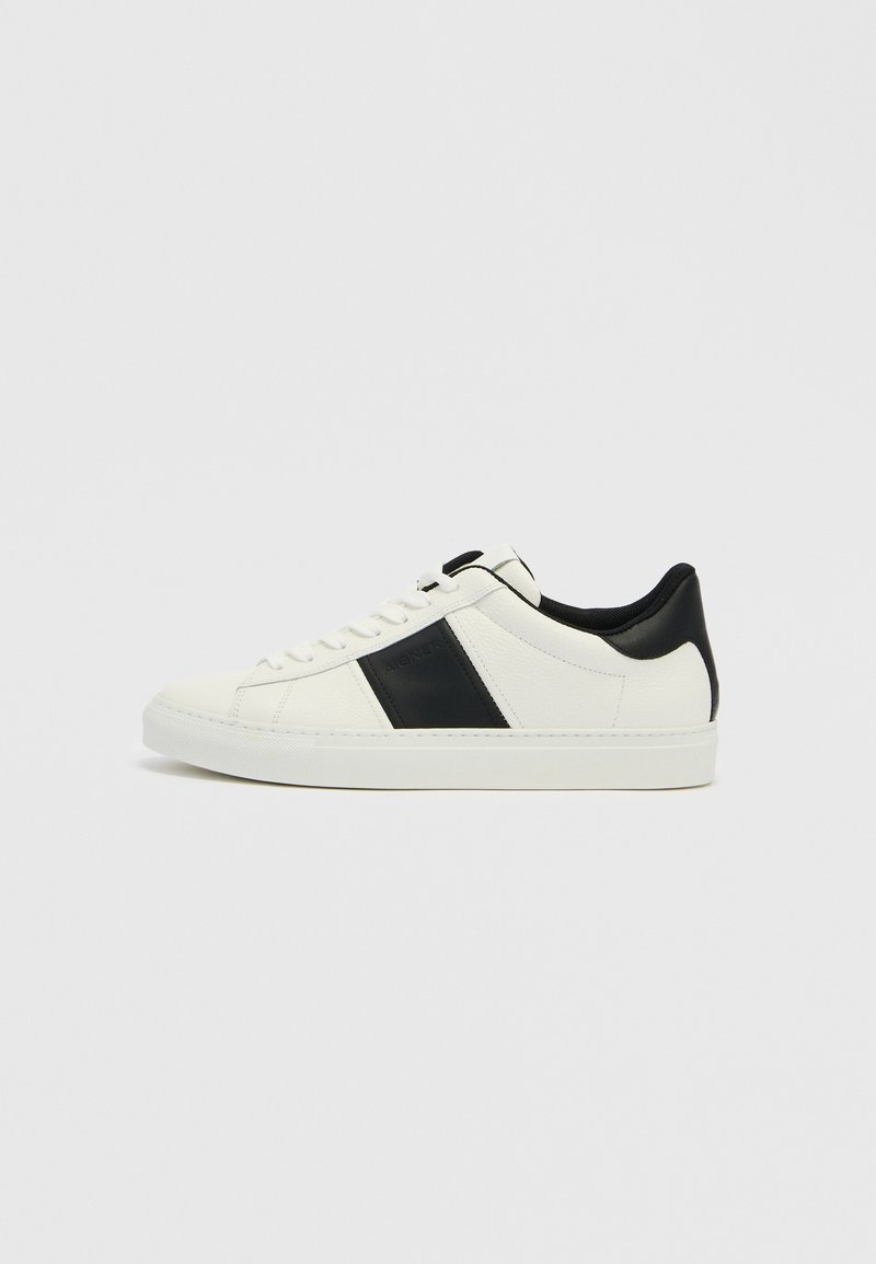 Weißer Sneaker mit niedrigem Schaft, schwarzer Ferse und seitlichem Streifen, Schnürverschluss und flacher Sohle vor schlichtem hellem Hintergrund.