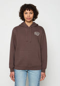 Person mit lockigem Haar, der einen braunen GAP-Hoodie mit Taschen vorne und eine blaue Jeans trägt, steht vor einem einfachen weißen Hintergrund.