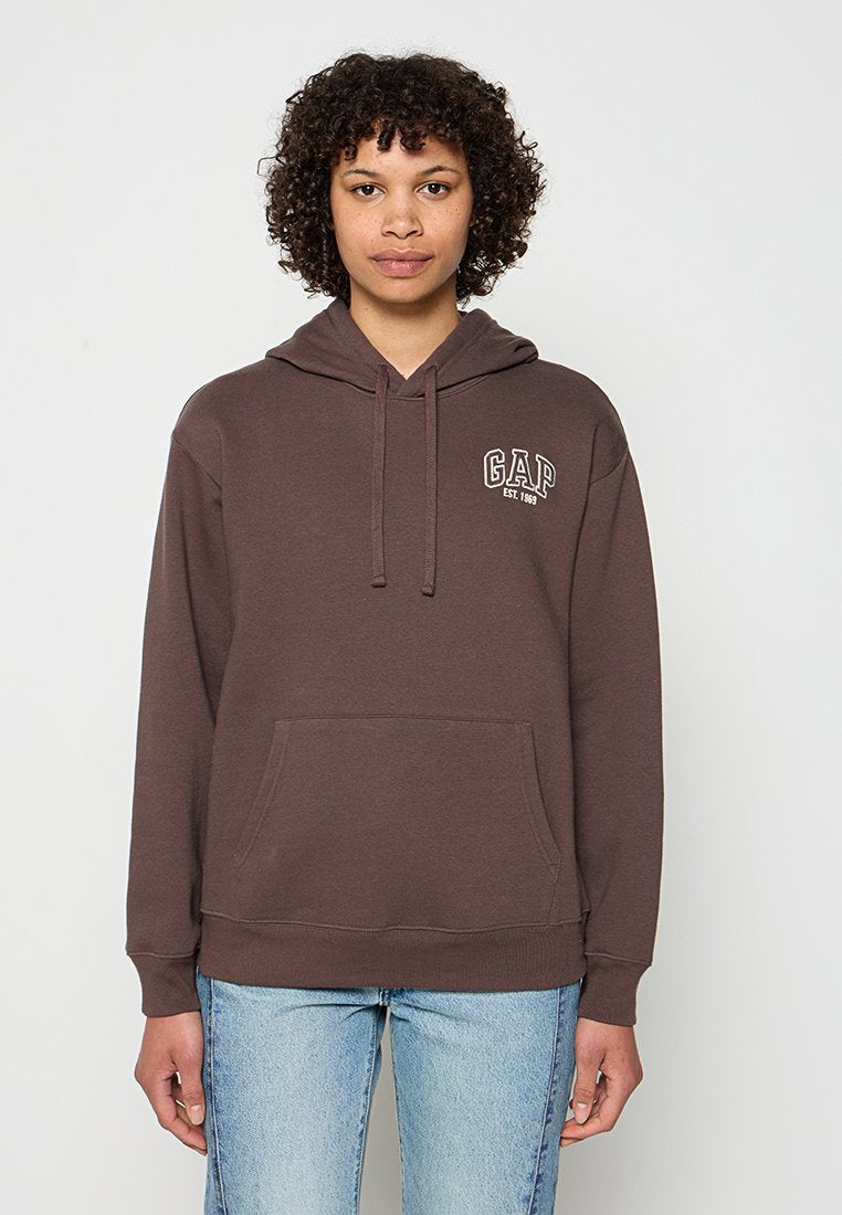 Person mit lockigem Haar, der einen braunen GAP-Hoodie mit Taschen vorne und eine blaue Jeans trägt, steht vor einem einfachen weißen Hintergrund.