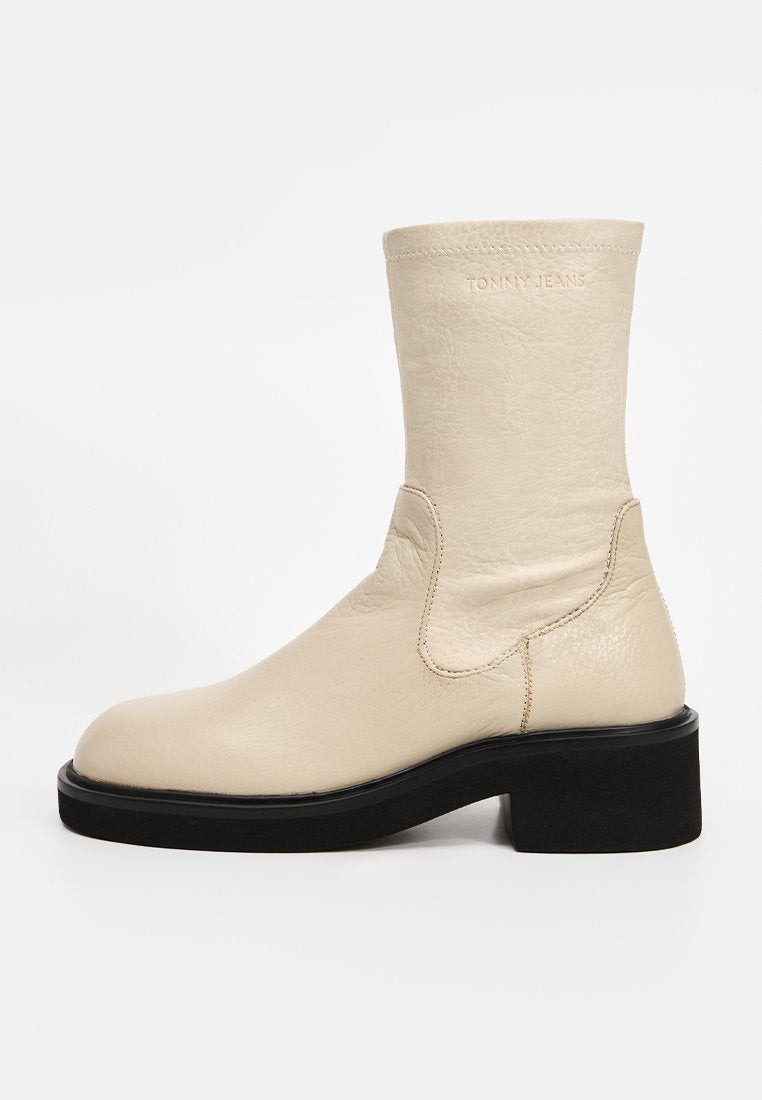 Cremefarbener Lederstiefel bis zur Mitte der Wade mit schwarzem, klobigem Absatz und Sohle, mit subtiler "TOMMY JEANS" Prägung nahe dem oberen Rand.