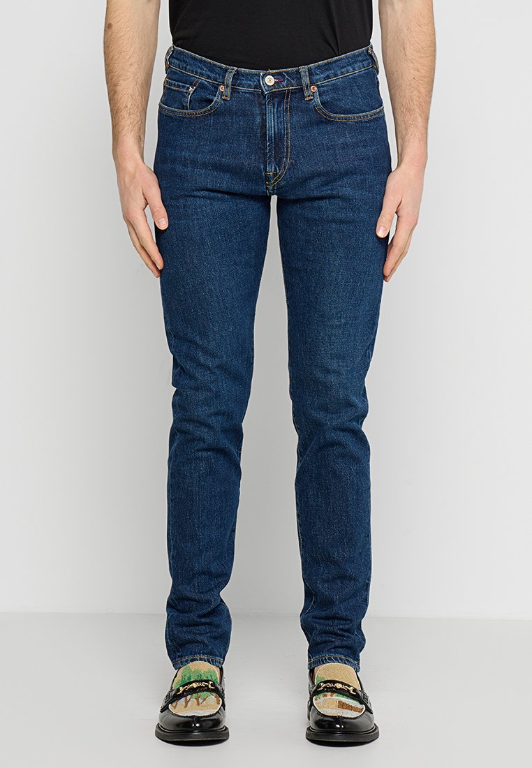 Dunkelblaue Jeans im Slim-Fit-Stil. Aus Denim gefertigt, mit fünf Taschen, einem Reißverschluss und gelben Nähten. Getragen mit schwarzen Slippern.