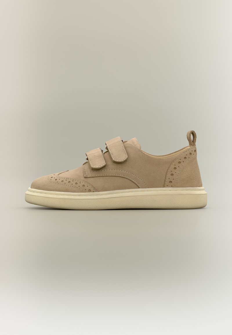 Beiger Wildleder-Sneaker mit doppelten Klettverschlüssen, perforierten dekorativen Mustern und einer cremefarbenen Gummisohle, seitlich auf hellem Hintergrund gezeigt.