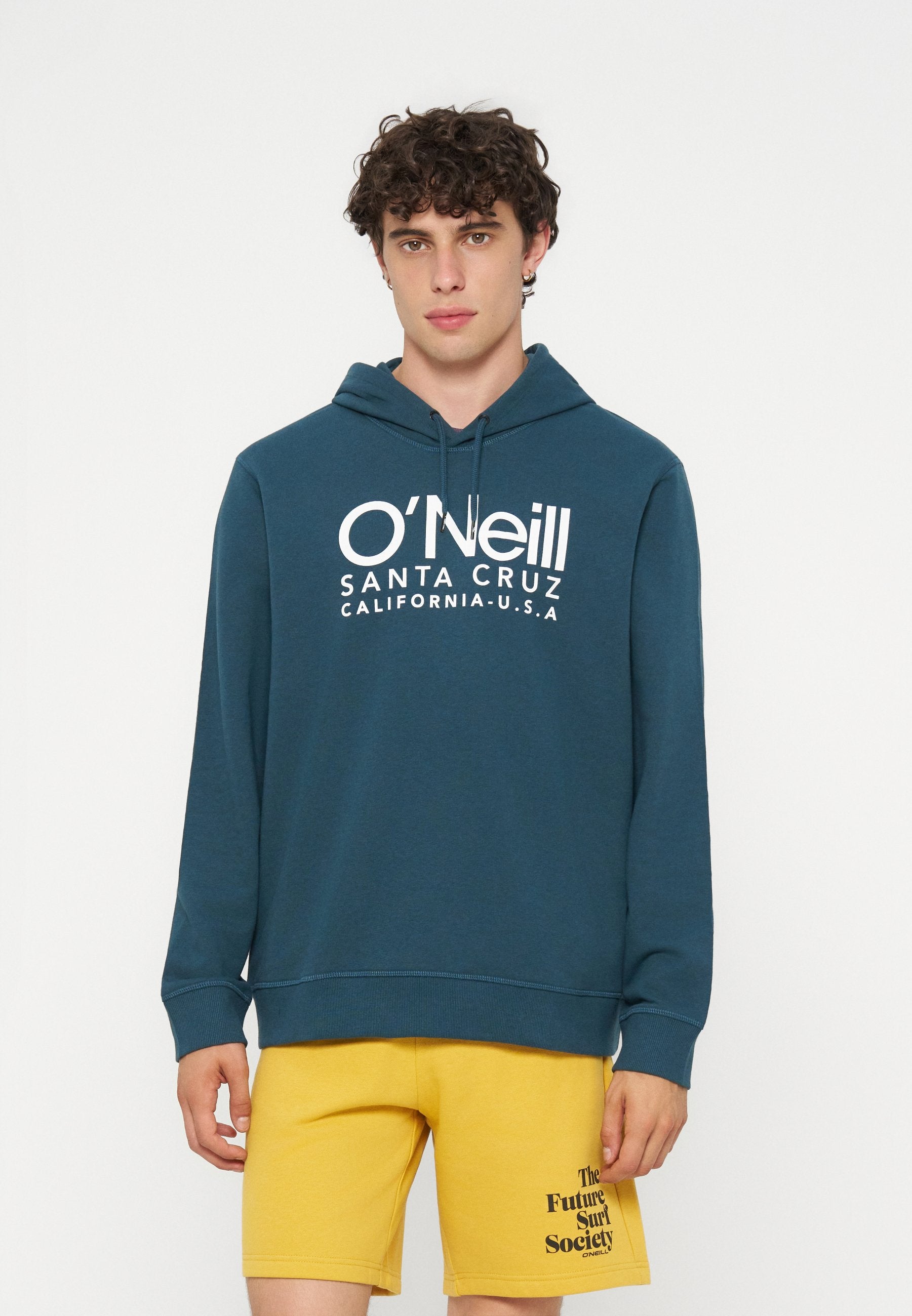 Junger Mann mit lockigem Haar, der einen blauen O'Neill Hoodie und gelbe Shorts mit dem Text "The Future Surf Society" am linken Bein trägt.