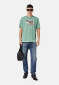 Mintgrünes T-Shirt mit kurzen Ärmeln und grafischem Druck, kombiniert mit blauen Jeans, schwarzen Lederschuhen und einer schwarzen Tasche. Sonnenbrille getragen.