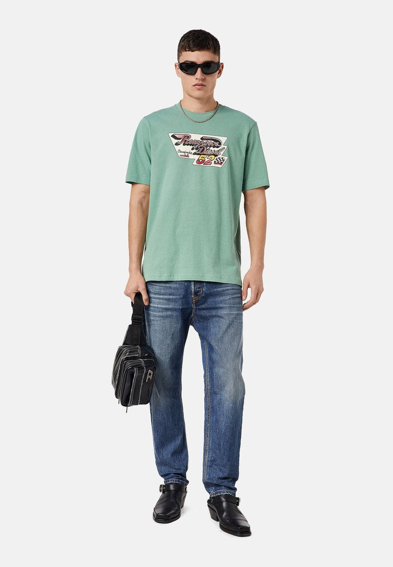 Mintgrünes T-Shirt mit kurzen Ärmeln und grafischem Druck, kombiniert mit blauen Jeans, schwarzen Lederschuhen und einer schwarzen Tasche. Sonnenbrille getragen.