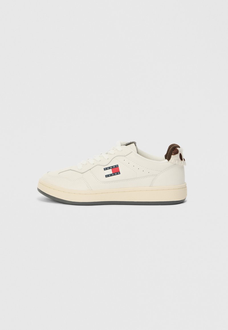 Weiße Sneaker mit einem glatten Lederoberteil, das mit einem Markenpatch in Rot, Weiß und Blau verziert ist, sowie einem strukturierten braunen Akzent an der Ferse.