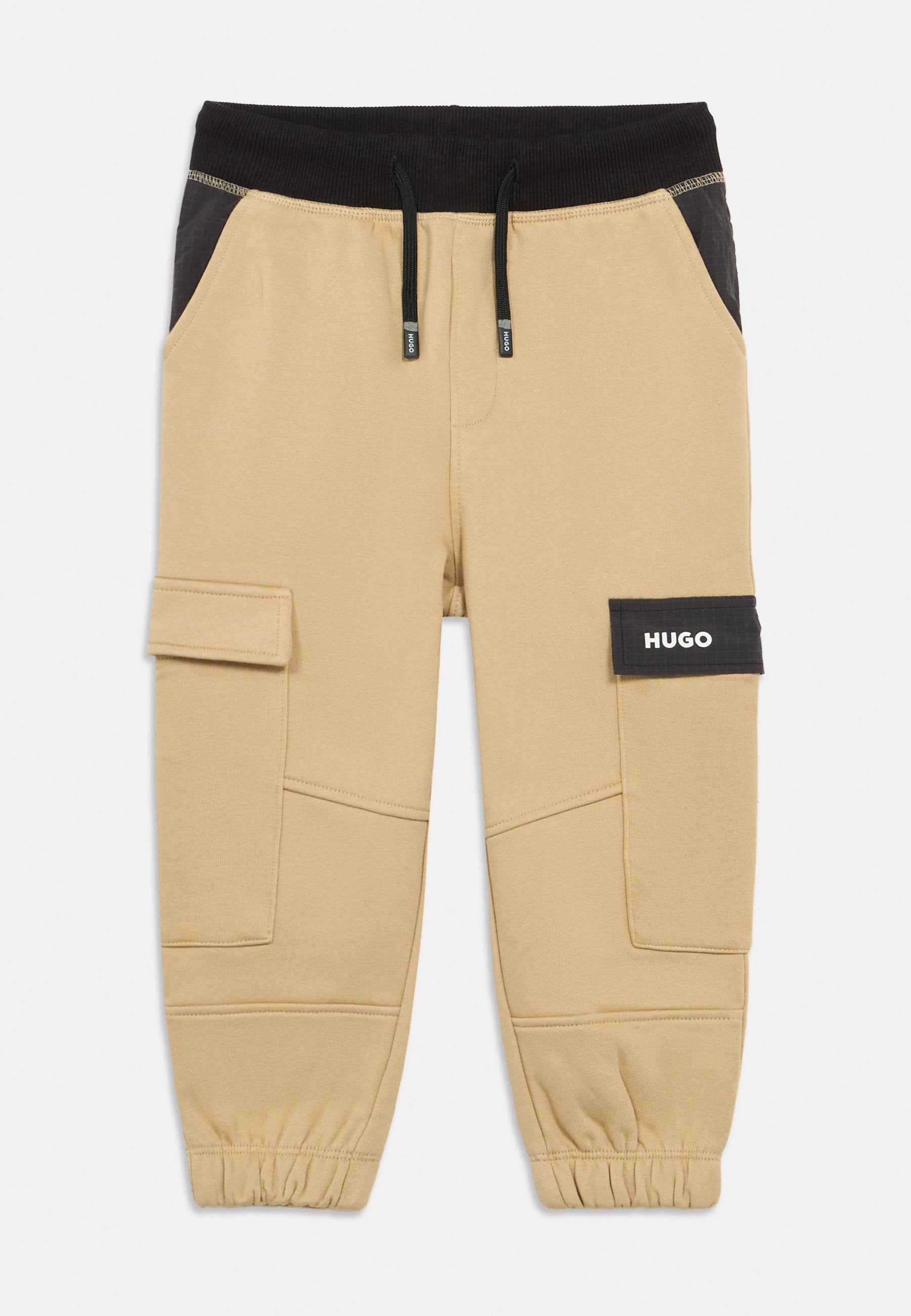 Beige Cargo-Hosen mit einem schwarzen Bund, der einen verstellbaren Kordelzug aufweist, seitlichen Taschen und einem auffälligen Patch mit der Aufschrift "HUGO". Elastische Bündchen an den Knöcheln.