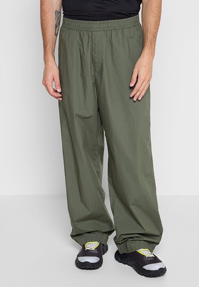 Herren-Hosen in Olivgrün aus leichtem Stoff; mit einem elastischen Bund und lockerer Passform. Kombiniert mit schwarzen Schuhen.