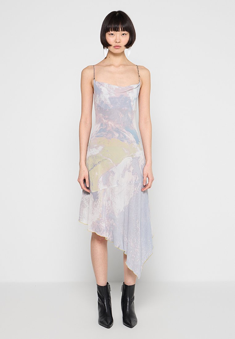 Frau trägt ein pastellfarbenes, asymmetrisches Slip-Kleid mit dünnen Trägern und schwarzen Stiefeletten, steht vor einem einfarbigen hellen Hintergrund.