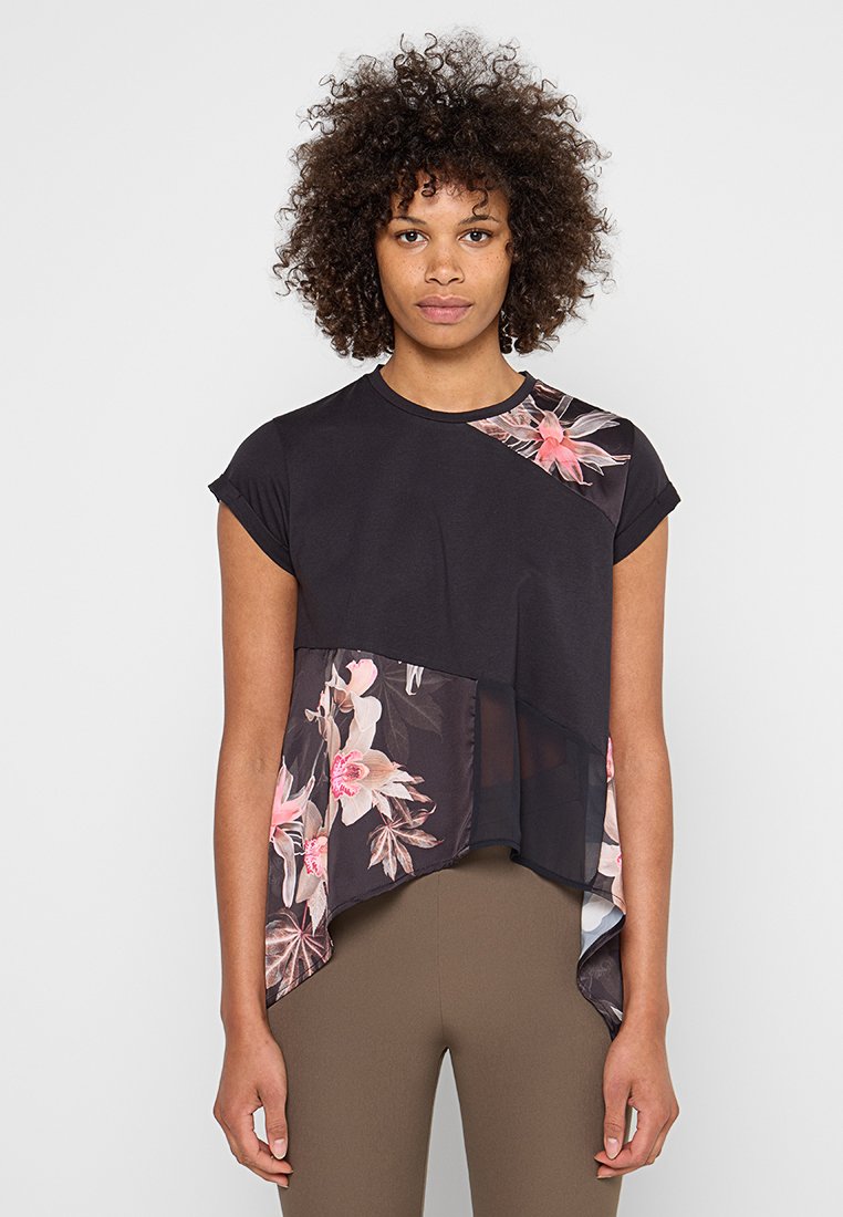 Frau mit lockigem Haar, die ein schwarzes, kurzärmeliges Oberteil mit rosa Blumenmustern und asymmetrischem Saum trägt, kombiniert mit braunen, eng anliegenden Hosen.