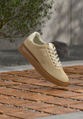 Beige Wildledersneaker mit weißen Schnürsenkeln und brauner Sohle, schwebend über einem Holzboden, im Hintergrund eine grüne Pflanze und eine graue Wand.